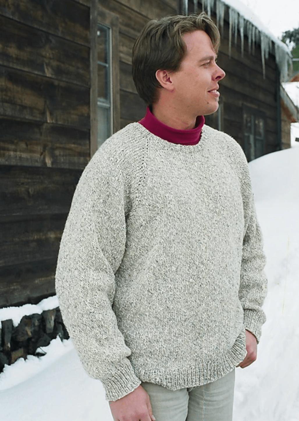 A man stands outside in snow by a rustic wooden building, wearing the Knitting Pure & Simple Neckdown Pullover for Men #991 over a burgundy turtleneck. He looks to the side with one hand in his pocket as icicles hang from the roof.