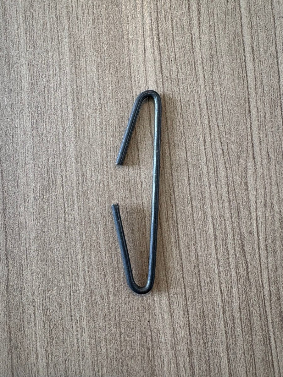 A Leclerc Loom Harness Hook A3 by Leclerc Looms, resembling a bent and broken metal paperclip with one end detached from the main loop, is lying on a wooden surface.