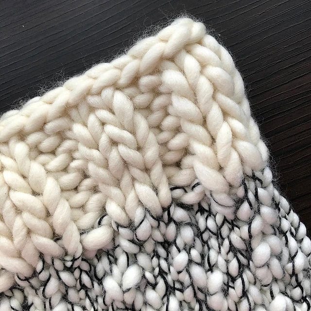 Close-up of the Aguas Blancas Cowl Pattern by Kristel Nieves (Ravelry Instore), featuring chunky, hand-knit cream and black Malabrigo merino wool with detailed stitches on a dark wooden surface.