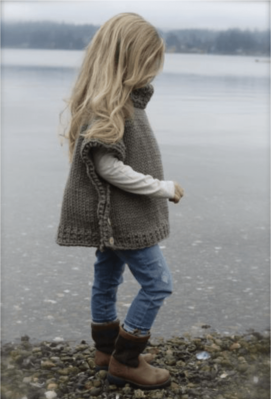 A child wearing an Azel Pullover Pattern by Heidi May knit from super bulky yarn stands by a lake, styled with a long-sleeved shirt, jeans, and boots.