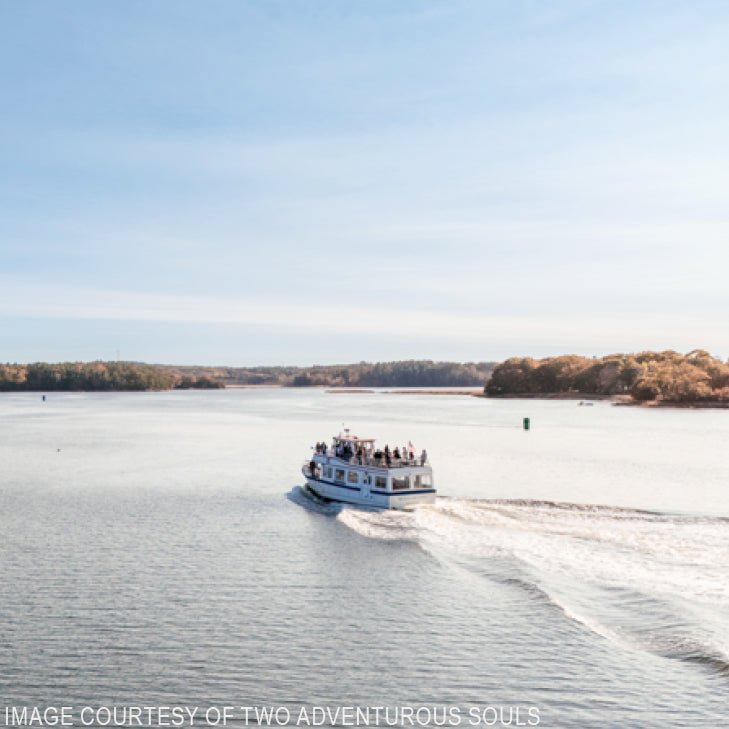 A white tour boat with passengers enjoys the Halcyon Yarn "Cast Off & Cast On: 2-Hour Kennebec Lighthouse & Nature Cruise (July 22)" on a calm river near the Maine Maritime Museum, with trees lining the shore under a clear blue sky.