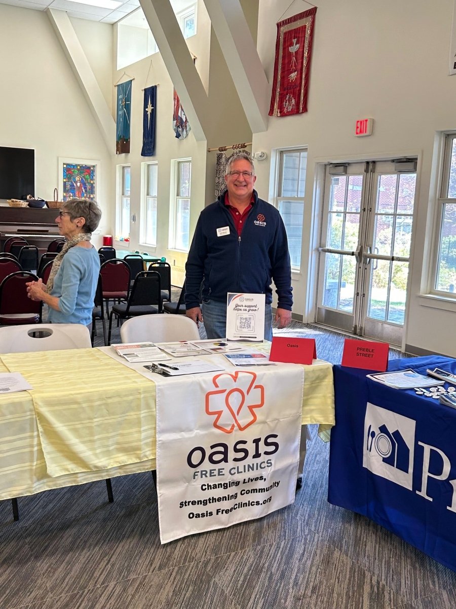 At a community center, a man in a red shirt and navy jacket stands behind an Oasis Free Clinics banner and table displaying info and handwoven scarves for the Halcyon Yarn Weaving Demo + Pop-Up Shop to benefit Oasis Free Clinic, May 8-9.