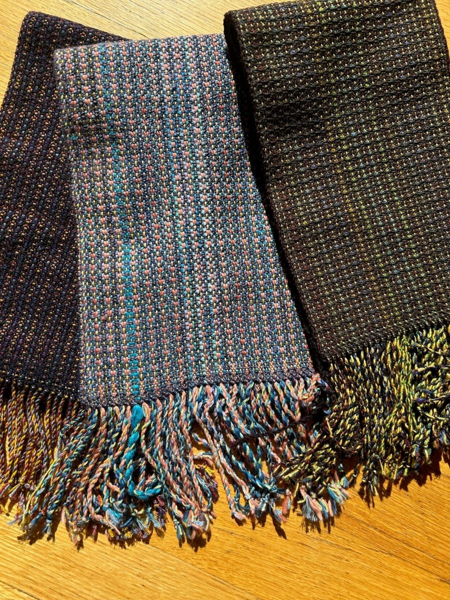 Three handwoven scarves by Halcyon Yarn—navy, blue/pink, and dark green/yellow—are displayed side by side in sunlight for the Weaving Demo + Pop-Up Shop, Oasis Free Clinic Benefit, May 8 & 9.
