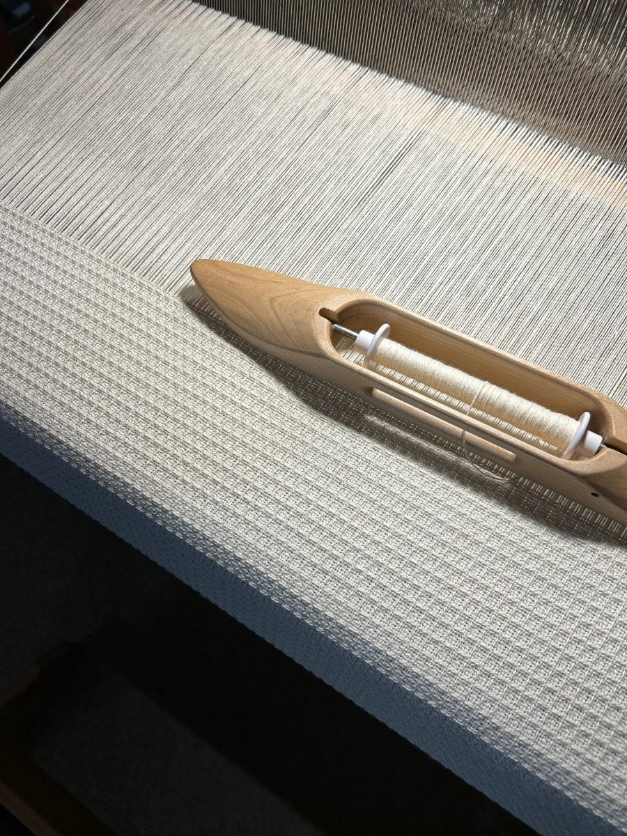 A close-up of a wooden weaving shuttle on a loom with fine white threads, taken at Halcyon Yarn’s Weaving Demo + Pop-Up Shop for Oasis Free Clinic Benefit, Fri & Sat May 8–9 (all day), where handwoven scarves are crafted.