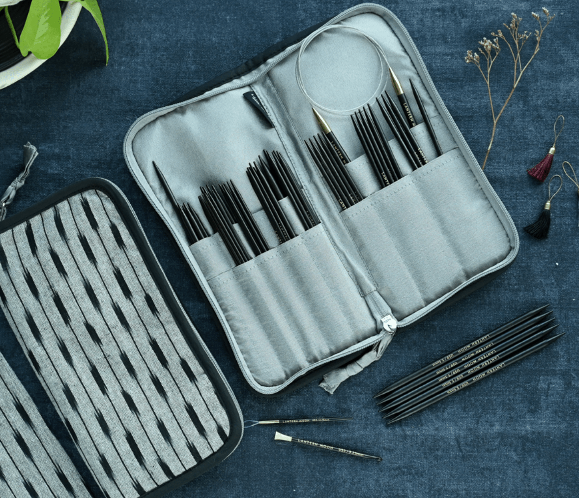 The Lantern Moon DPN Needle Case by Bryson Distributing, Inc. is shown open, neatly holding knitting needles and accessories, displayed on dark blue IKAT fabric with dried flowers and a nearby plant for a cozy touch.