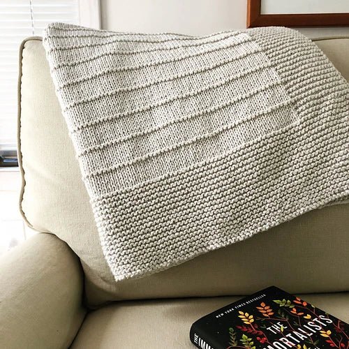 The On The Porch Baby Blanket Pattern by Fifty Four Ten Studio (Ravelry Instore) is shown in cream, featuring modern horizontal textured stripes, draped over a light beige armchair beside a book with a colorful cover.