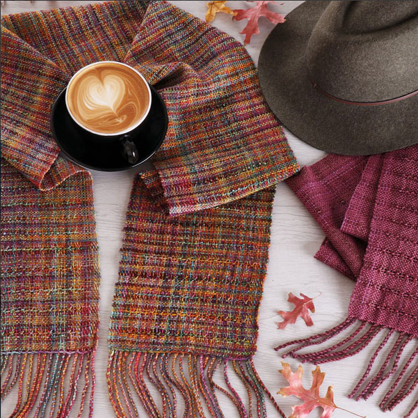 A latte with heart-shaped foam rests on a colorful woven scarf. Nearby are a gray hat, pink scarf, autumn leaves, and Ashford Handicrafts Limited's "Rigid Heddle Weaving Basics And Beyond" by Deborah Jarchow on a light wooden surface.