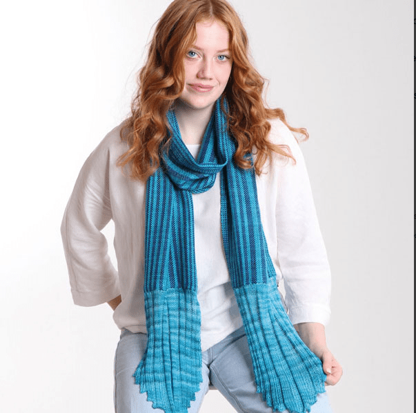 A woman with long, wavy red hair models a blue knitted scarf and casual outfit, showcasing effortless style inspired by "Rigid Heddle Weaving Basics And Beyond" by Deborah Jarchow (Ashford Handicrafts Limited), against a plain white background.