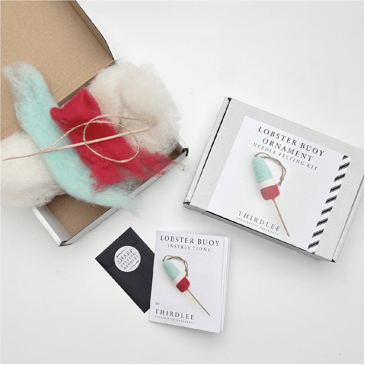 The Thirdlee Maine Needle Felting Kits Lobster Buoy by Thirdlee Co includes colored wool, twine, a felting needle, and instructions to make a buoy ornament. Items are displayed beside an open and closed kit box on a white surface.