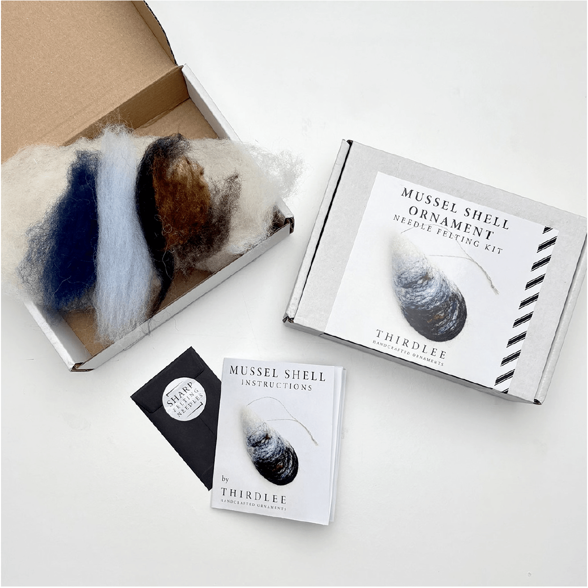 The Thirdlee Co Thirdlee Maine Needle Felting Kits Mussel Shells is displayed with multicolored wool, a box featuring a mussel shell, a "Mussel Shell Instructions" booklet, and a pack of sewing needles.