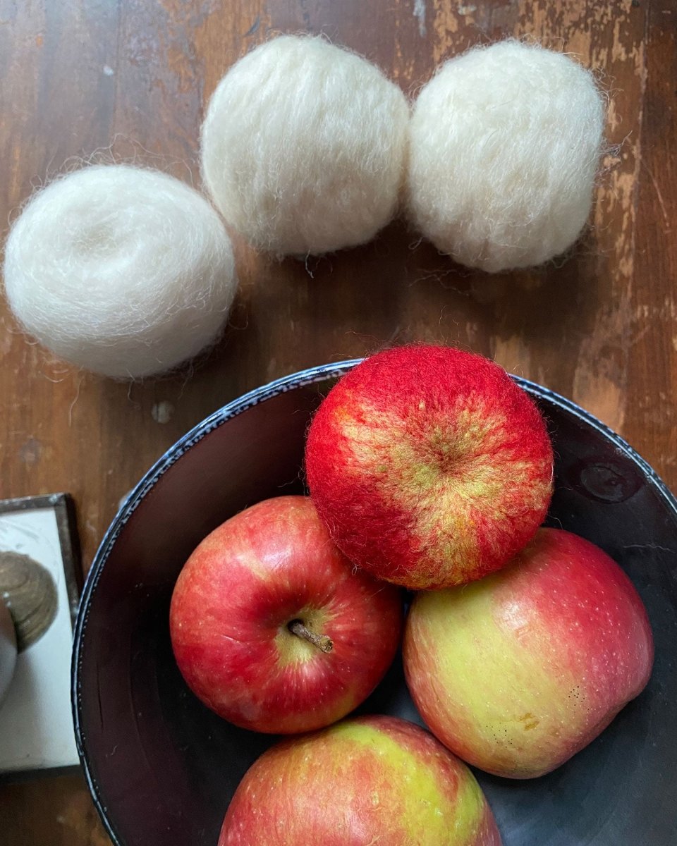 A black bowl containing four felted red apples rests on a wooden surface, evoking the creations from Halcyon Yarn’s “Needle Felting Class: Apples with Michelle Provençal (September 12th).” Above the bowl, three white wool dryer balls are neatly arranged in a row.