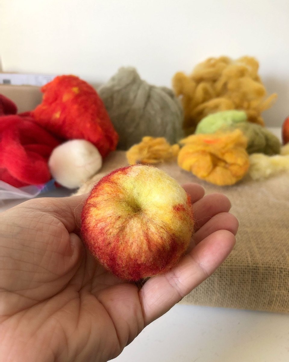A hand holds a small, felted wool apple in vibrant red and yellow tones. In the background, bundles of colorful wool roving and a piece of burlap rest on the table—perfect inspiration for those attending the Halcyon Yarn “Needle Felting Class: Apples with Michelle Provençal (September 12th)” or anyone eager to craft their own felted wool apples.