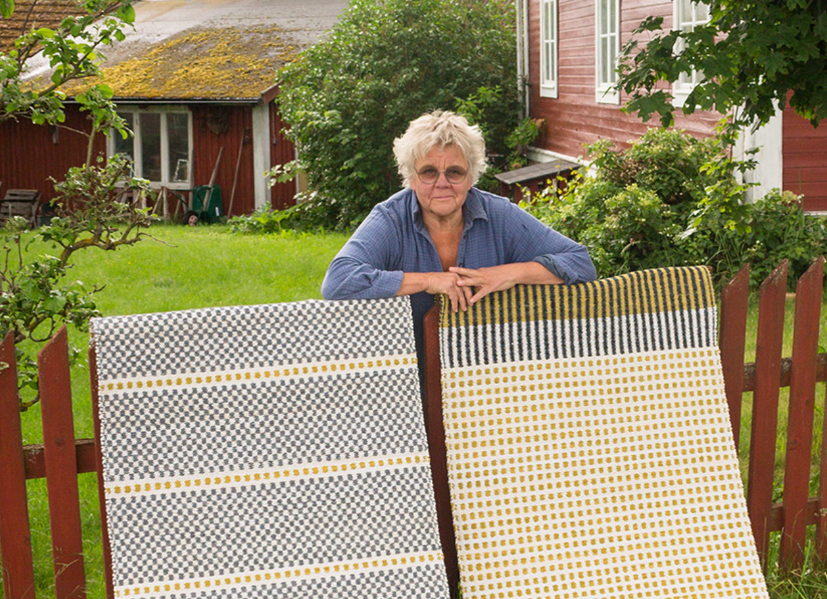 A woman leans on a wooden fence holding two geometric yellow and grey blankets inspired by Scandinavian traditions, featured in VÄV Bookazine 2026 by VÄV Magazine.