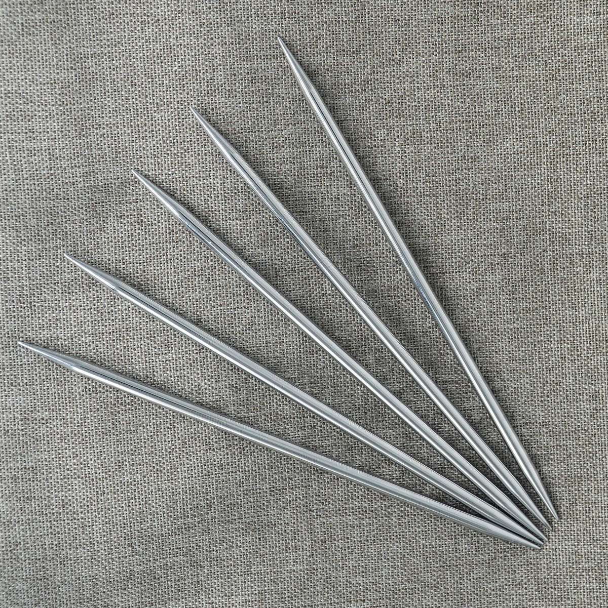 Six Knit Pro Nova Metal 6-inch double-point knitting needles from Accessories Unlimited are displayed in a fanned-out pattern on textured gray fabric.