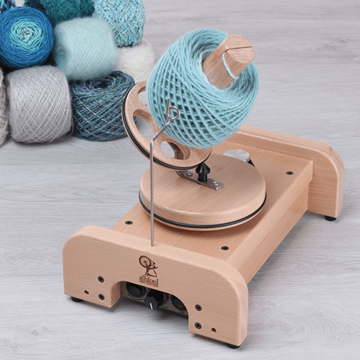 The Ashford Wooden Ball Winder by Ashford Handicrafts Limited holds a large spool of light blue yarn, with several colorful yarn balls arranged in the background on a light wood floor.
