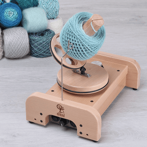 The Ashford Wooden Ball Winder by Ashford Handicrafts Limited holds a large spool of light blue yarn, with several colorful yarn balls arranged in the background on a light wood floor.