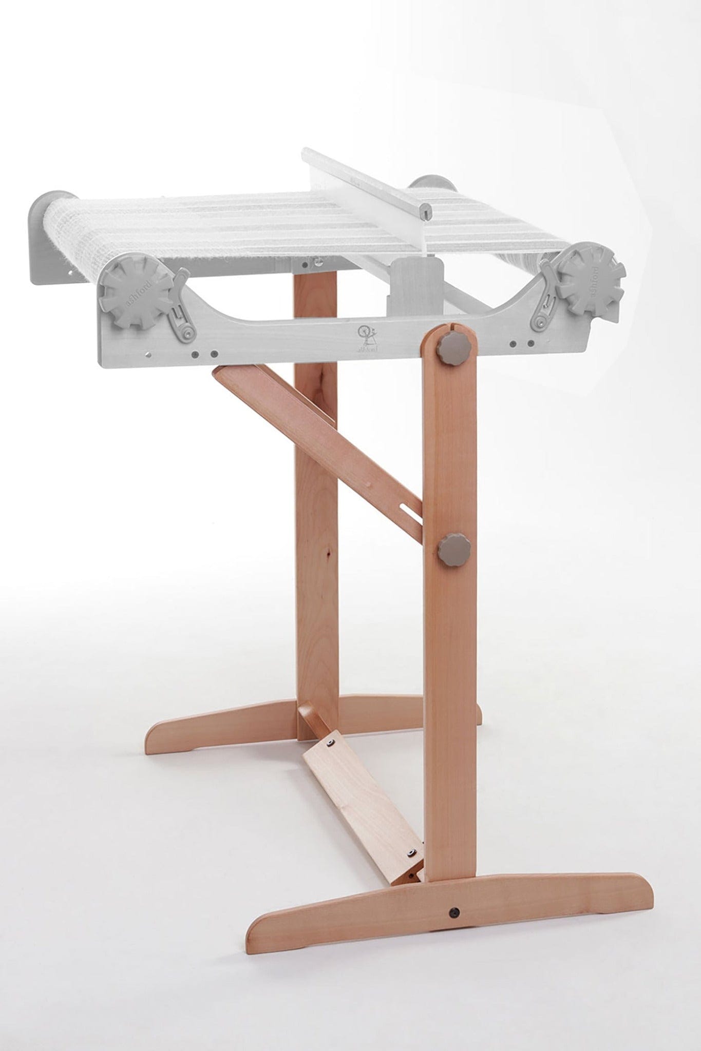 The Ashford Handicrafts Limited Ashford Rigid Heddle Floor Stand features a sturdy wooden base, adjustable ergonomic knobs, and a sleek design for comfortable weaving. White threads stretch across its top frame for easy setup.