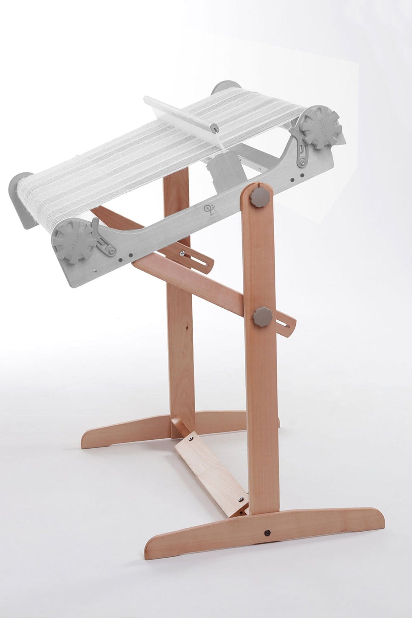 The Ashford Handicrafts Limited Ashford Rigid Heddle Floor Stand, featuring adjustable height and weaving angle, securely supports a rigid heddle loom with white threads against a plain, light-colored background.