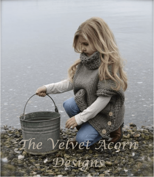 A girl wearing a chunky, super bulky yarn Azel Pullover Pattern by Heidi May turtleneck poncho squats by the water’s edge holding a metal bucket.