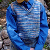 A child models the Basic Vest for Children, #256 by Bryson Distributing, Inc.—a blue and multicolored worsted weight sweater vest—over a blue button-up shirt outdoors near rocks, with only the torso and lower face shown.