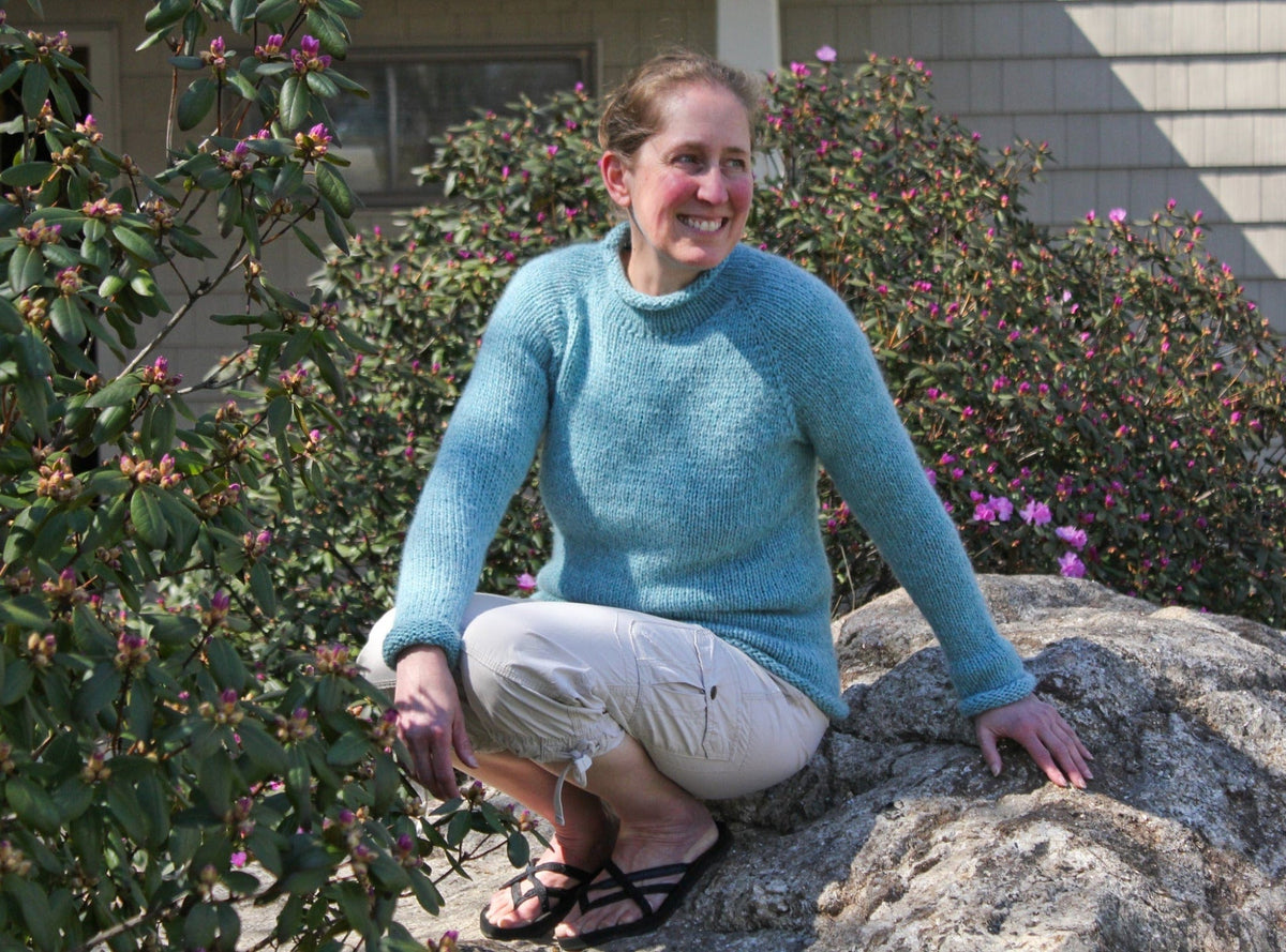 A woman wearing the Knitting Pure & Simple Bulky Neck Down Pullover #992 in blue and light pants smiles as she sits on a large rock, surrounded by blooming bushes with pink flowers in a sunlit outdoor setting. The cozy texture and delicate knitting pattern of the pullover add charm to the vibrant scene.