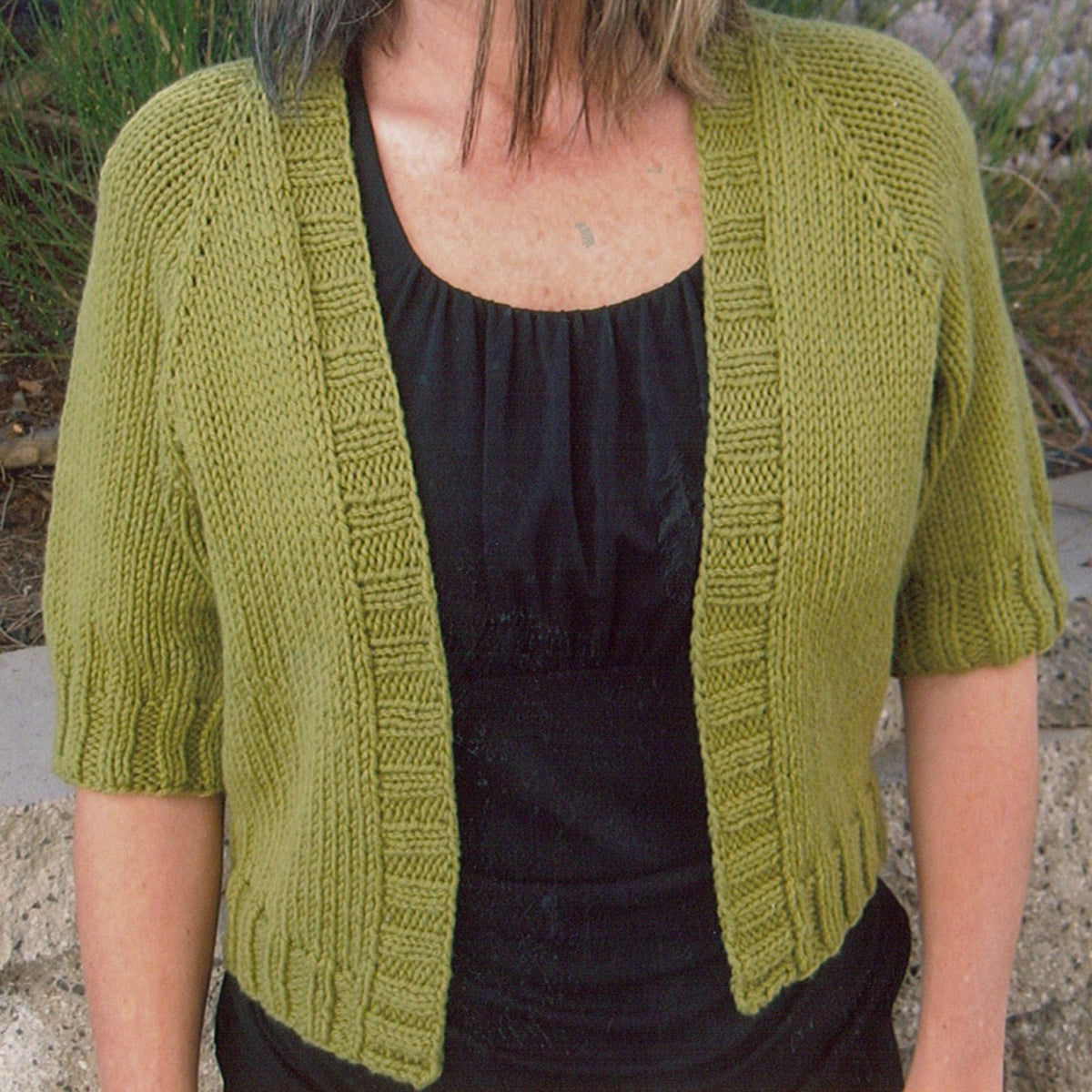 A person wears the Summer Open Cardigan by Knitting Pure & Simple—a short-sleeved, open green knit in worsted weight—over a black top, shown from shoulders to waist outdoors. Download available.