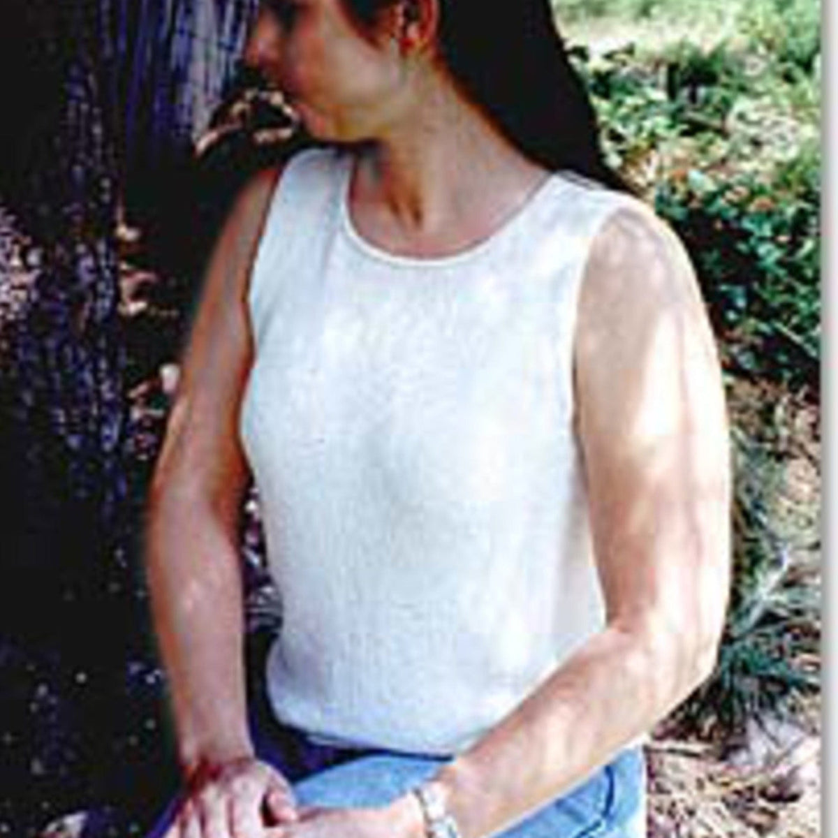 A woman with long dark hair wears the Knitting Pure & Simple Basic Shell Tank Top Pattern (#997) in white and blue jeans, sitting outdoors on a bench with trees and greenery behind her, looking left with her hands on her lap.