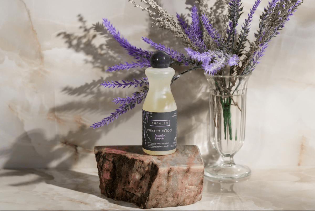 A 100 ml (3.3 oz) bottle of Eucalan Wool Wash by Eucalan, a lanolin-enriched and biodegradable textile cleanser, rests on a stone slab with a glass vase of lavender flowers behind it, set against a light marble background.