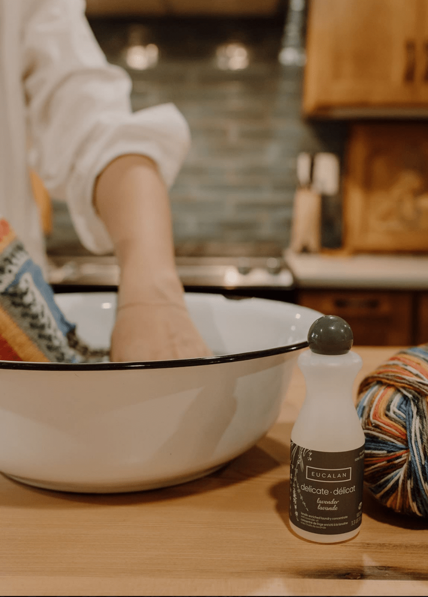 In a cozy kitchen, a person hand-washes a vibrant knitted piece in a white basin using Eucalan Wool Wash 100 ml / 3.3 oz Bottle by Eucalan—a lanolin-enriched, biodegradable no-rinse cleanser—while a yarn ball and the bottle rest on a wooden table.