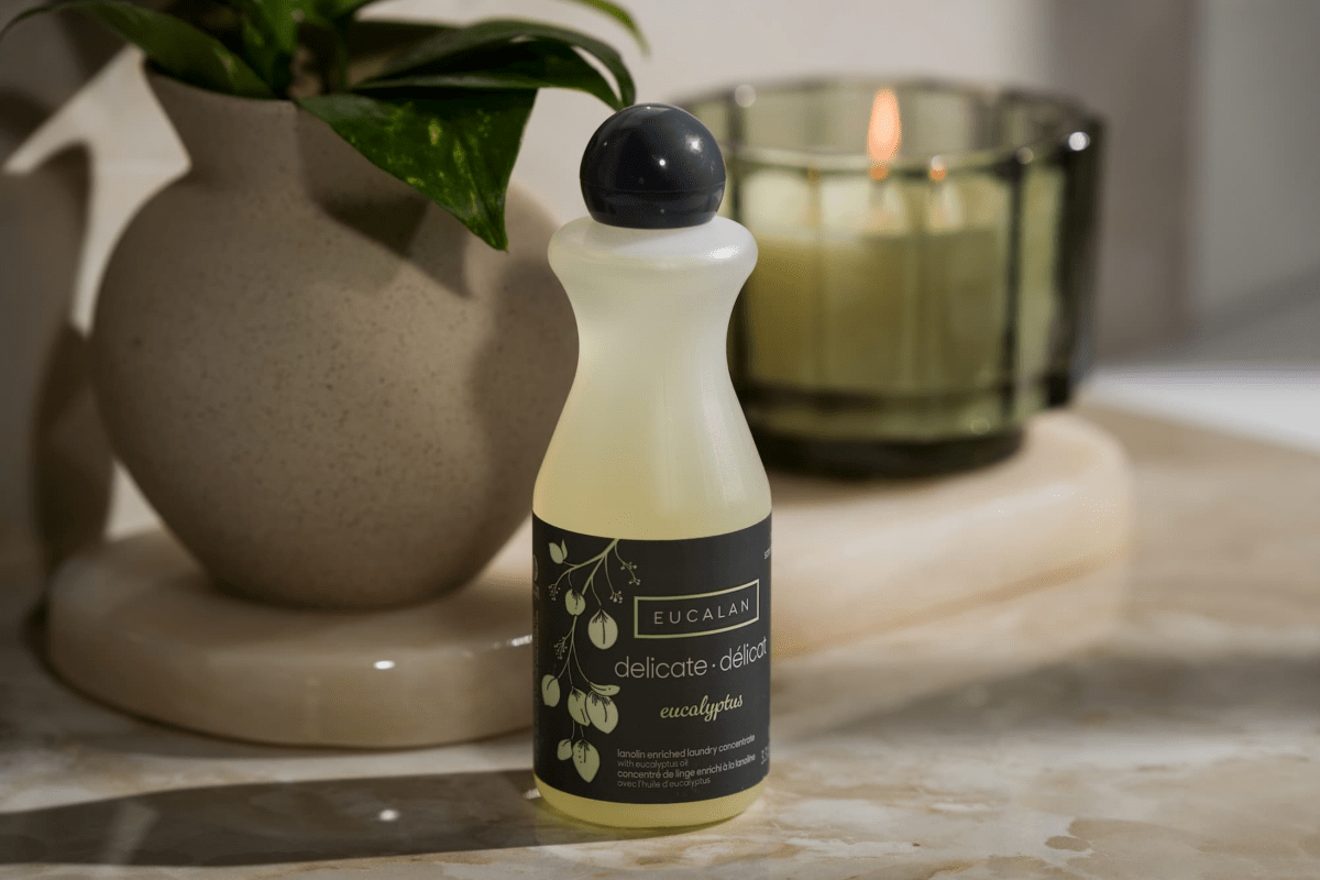 A 100 ml (3.3 oz) bottle of Eucalan Wool Wash by Eucalan, a biodegradable lanolin-enriched cleanser that requires no rinsing, sits on a marble surface beside a green glass candle and a beige vase holding a green plant, all softly illuminated.
