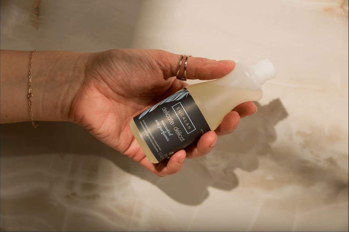 A hand with gold rings holds a small, open bottle of Eucalan Wool Wash 100 ml / 3.3 oz from Eucalan, set against a light marble background.