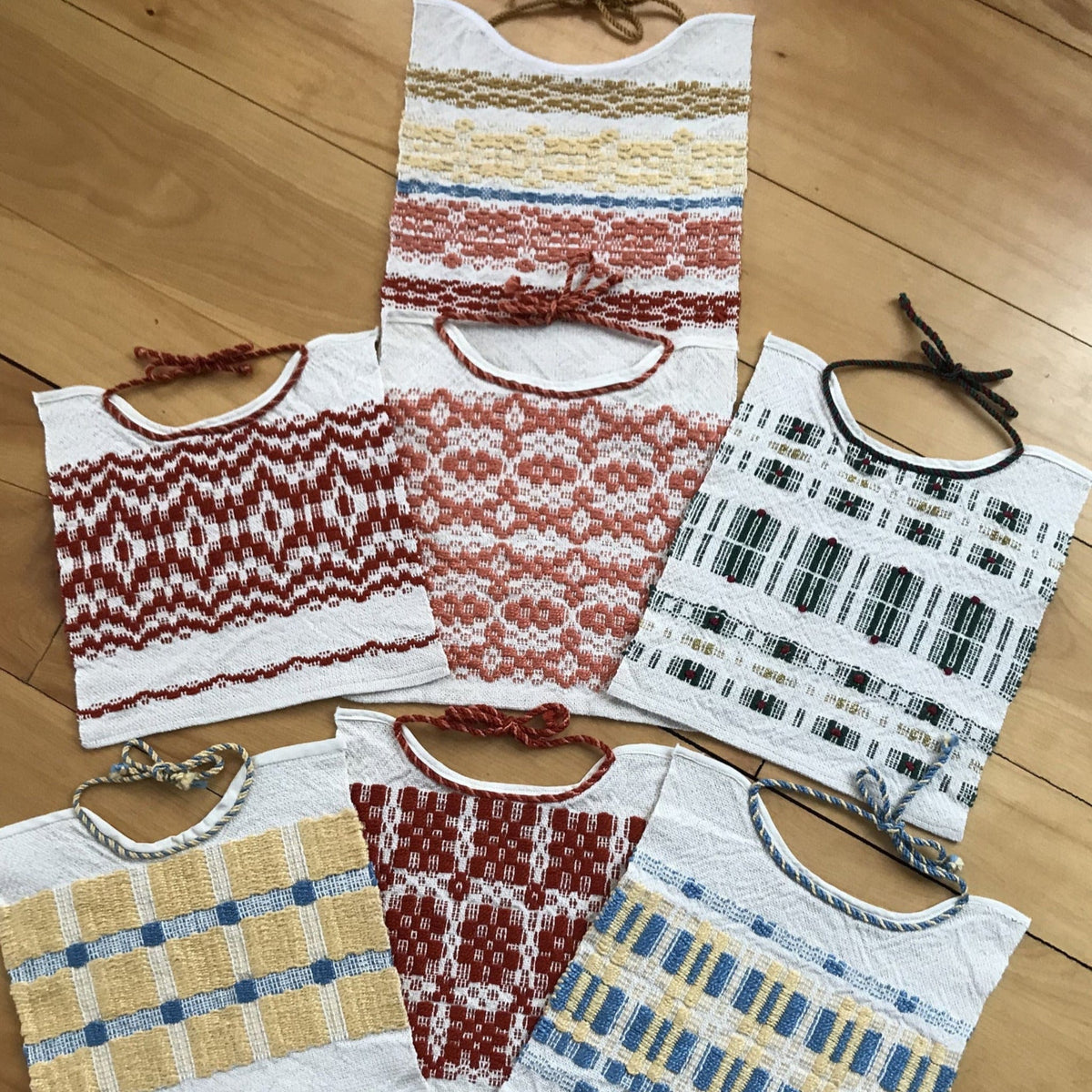 Six Show Off Baby Bib Woven Patterns by Halcyon Yarn, each with geometric designs in red, yellow, blue, and green, are displayed on a light wooden floor. Braided neck ties add charm to this creative stash buster project.
