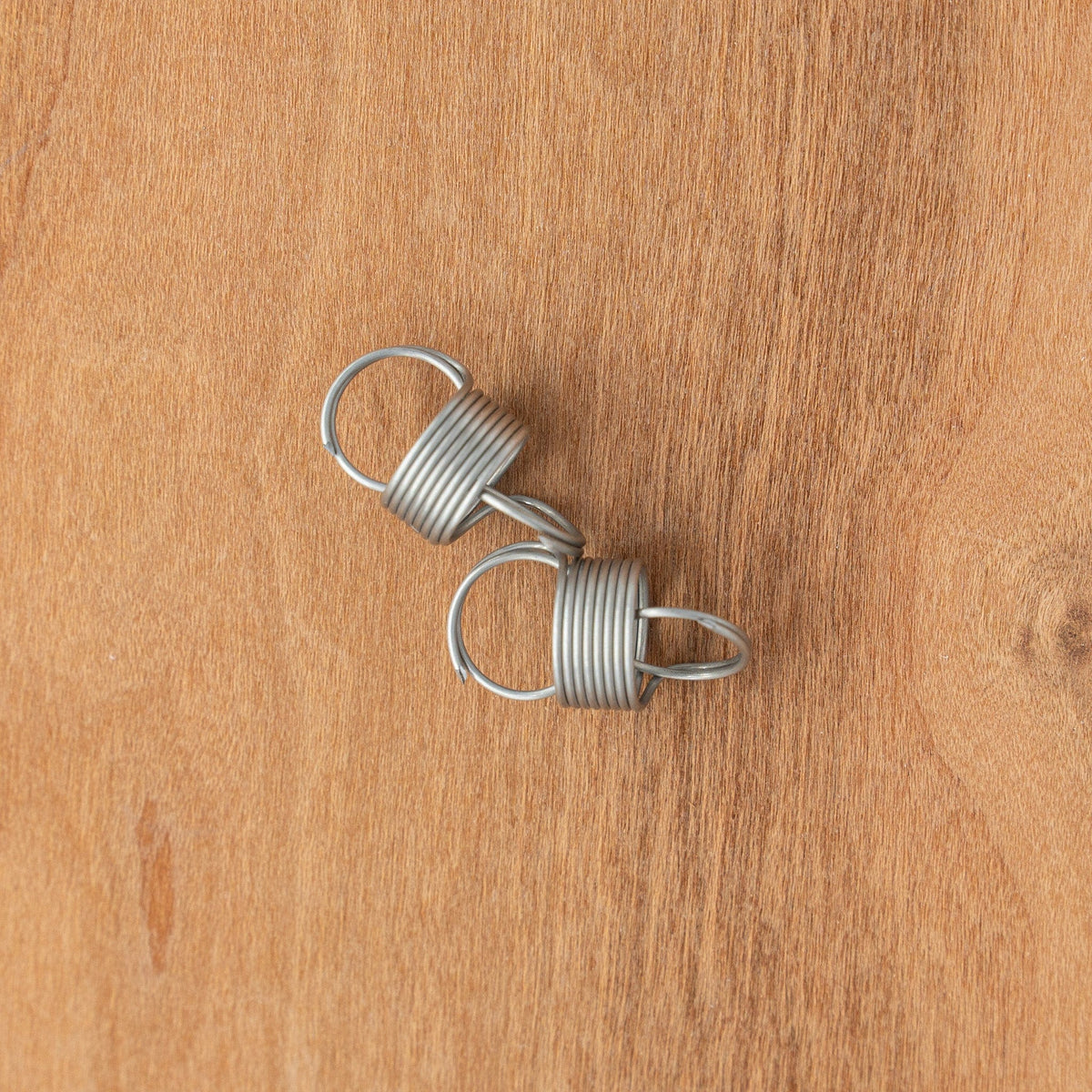 A Kromski Spinning Wheel Spring by Kromski North America lies on a light brown wooden surface. The metal spring, sold individually, features loops at each end and resembles a traditional spinning wheel spring.