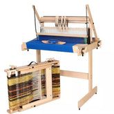 A Louët Table Loom by Louet Inc., crafted from solid beech hardwood, features a blue weaving area with multiple heddles. Below it, a sturdy frame displays partly completed yellow, brown, and black fabric.