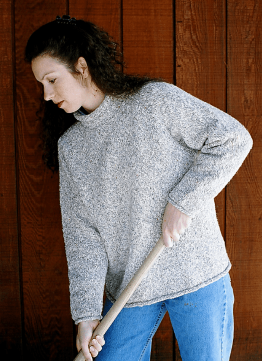 A woman with long curly hair wearing the Knitting Pure & Simple Neckdown Pullover for Women #9724 and blue jeans sweeps the floor with a broom in front of a wooden wall.