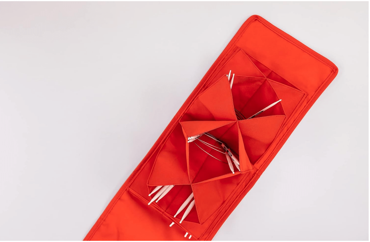 A Bryson Distributing, Inc. Circular Knitting Needle Case, red and rectangular with multiple compartments, is shown from above on a light background holding several thin white and red knitting needles.