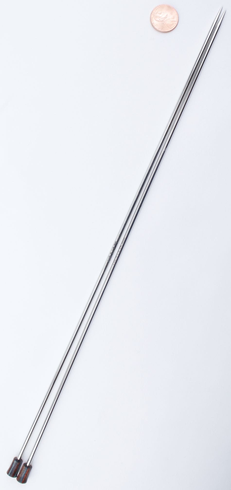 Two Knit Pro Nova Metal Single Point Knitting Needles (10" or 14") by Accessories Unlimited, with dark ends, lie parallel on a white background with a penny above them for size comparison.