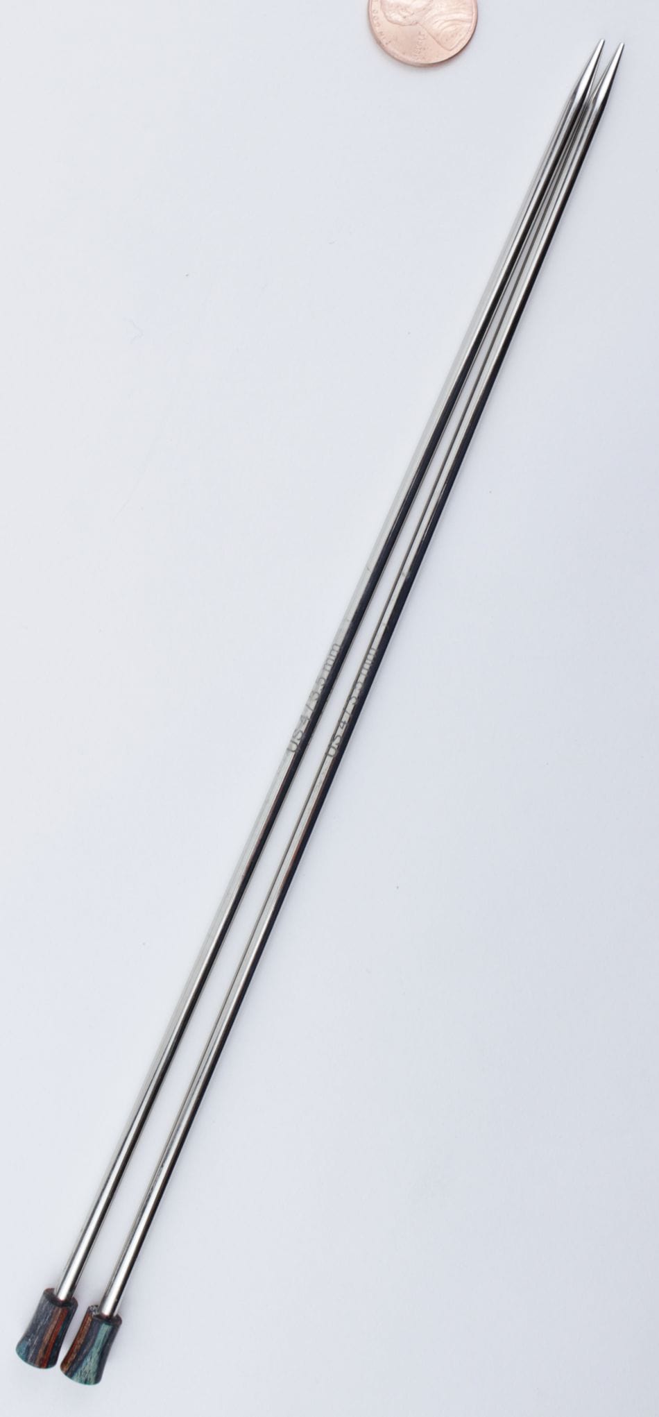 Two long Knit Pro (Knitters Pride) Nova Metal Single Point Knitting Needles, 10" or 14", from Accessories Unlimited, with flat colored ends, are placed parallel on a white surface. A coin near the top shows their scale.
