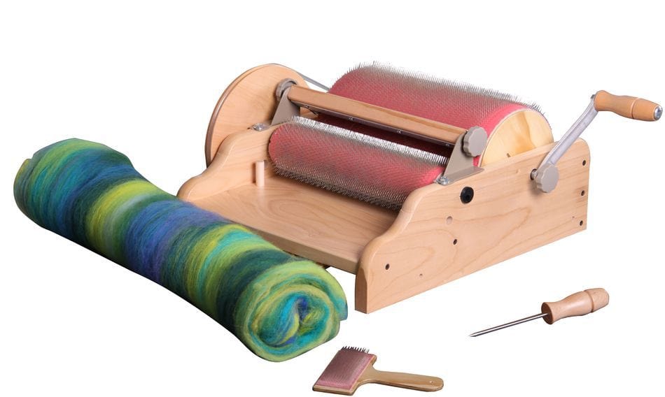 The Ashford Drum Carder by Ashford Handicrafts Limited is shown beside a rolled-up blend of blue, green, and yellow fiber. In front, a hand carder brush and a wooden-handled awl are positioned near the machine.