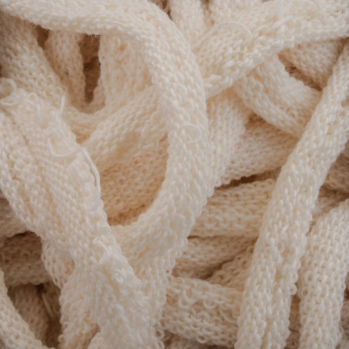 Close-up of intertwined, soft, off-white Harrisville Potholder Loops by Friendly Loom. The texture appears cozy and slightly fuzzy, reminiscent of intricate knit patterns from a traditional loom. The loops create an abstract, woven appearance with a sense of warmth and comfort.