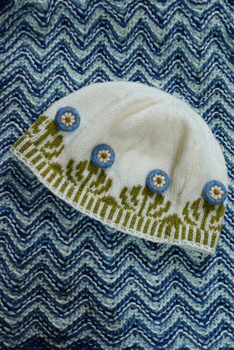 A cozy white knitted hat adorned with a green pattern and four blue Dorset Buttons—crafted using traditional button-making techniques taught in "Discover Dorset Buttons: A Hands-On Workshop with Linda Perry (October 24, 2026)" by Halcyon Yarn—is displayed on a blue and white zigzag patterned knitted background.