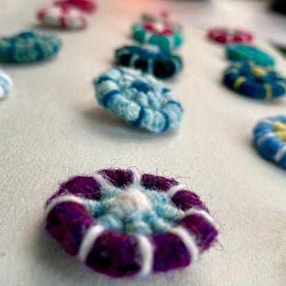 A close-up view of vibrant, round woven buttons or discs in dominant shades of purple, blue, and teal are beautifully arranged on a white surface. Inspired by traditional techniques featured in Dorset Buttons and reflecting the artistry taught in "Discover Dorset Buttons: A Hands-On Workshop with Linda Perry (October 24, 2026)" from Halcyon Yarn, the foreground is sharp and detailed while the background remains softly blurred.