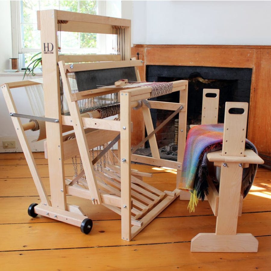 A wooden floor loom and stool setup is situated in a sunlit room. The loom holds a partially woven multicolored fabric. The equipment appears to be made from light-colored wood, and there's a window in the background letting in natural light.