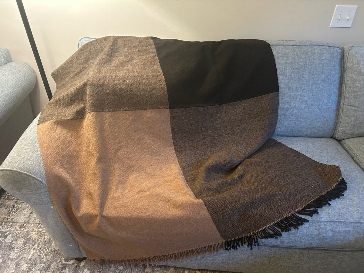 A color-blocked throw from Halcyon Yarn’s Weaving Demo + Pop-Up Shop (May 8-9, Oasis Free Clinic Benefit) in brown and black is draped over a light gray couch, with a decorative rug partially visible on the floor.