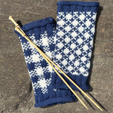 A pair of blue and white patterned, two-stranded fingerless gloves made with Halcyon Yarn appears on a stone surface, alongside two wooden needles. Inspired by the Cast Off & Cast On: 3-Hour Kennebec Lighthouse & Nature Cruise (August 11).