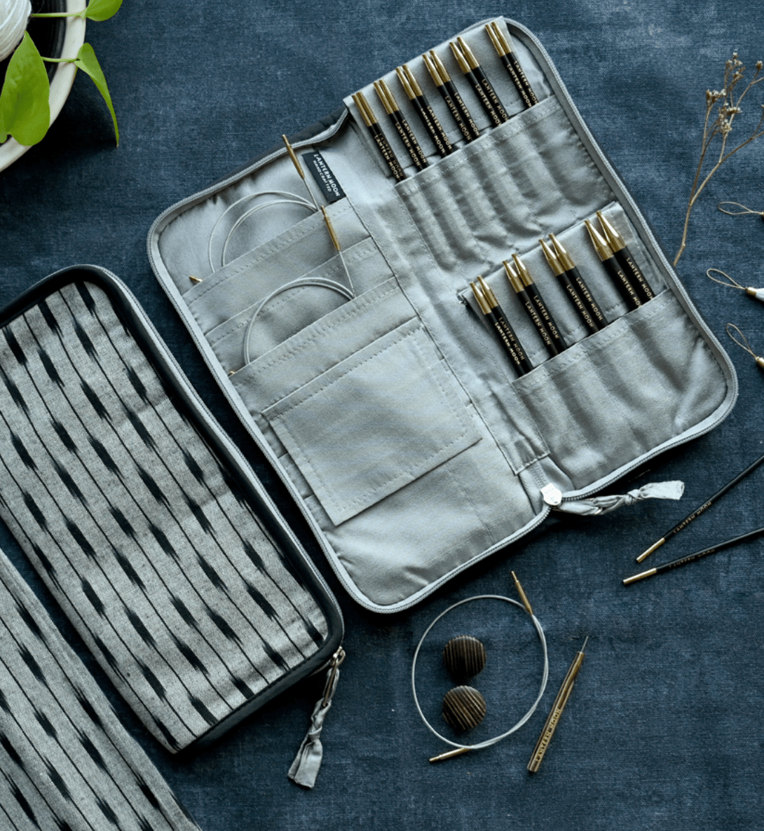 An open handcrafted Lantern Moon Interchangeable Needle Case by Bryson Distributing, Inc. sits on a blue surface, neatly organizing knitting needles, crochet hooks, and cables. Nearby are a closed matching case, yarn, and accessories.