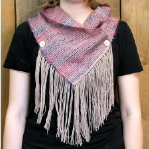 Woman wearing a black shirt and a triangular woven scarf featuring pink, purple, and gray hues. The scarf is fastened with two buttons and has long, beige fringe hanging from the bottom. The background consists of wooden panels.