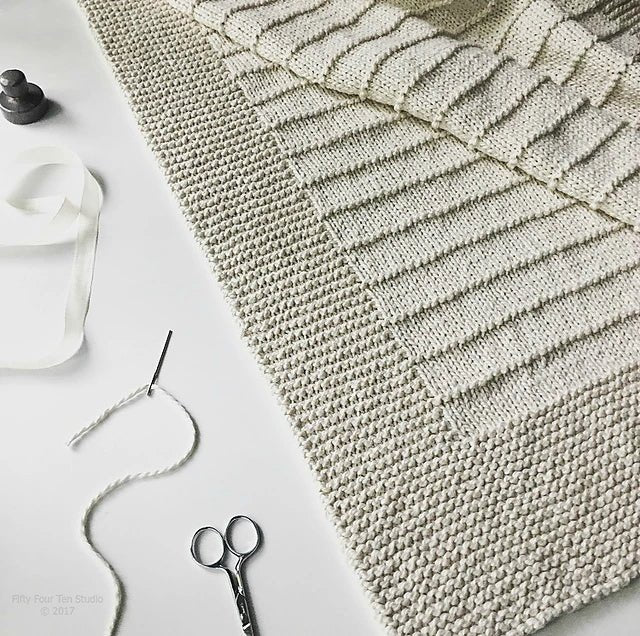 The On The Porch Baby Blanket Pattern by Fifty Four Ten Studio (print) from Ravelry Instore is shown in cream with textured borders and stripes, laid on white near a ribbon, scissors, needle with yarn, and wooden accents.