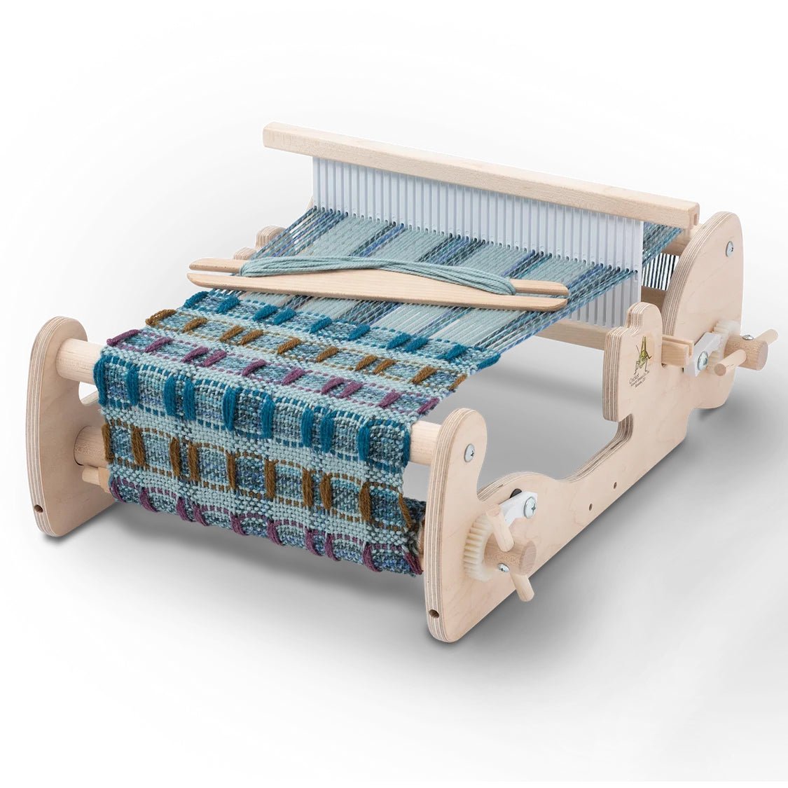 A Schacht Spindle Co. Cricket Rigid Heddle Loom sits on a tabletop with a partially woven blue, green, and brown checkered fabric, while a wooden shuttle rests on top.