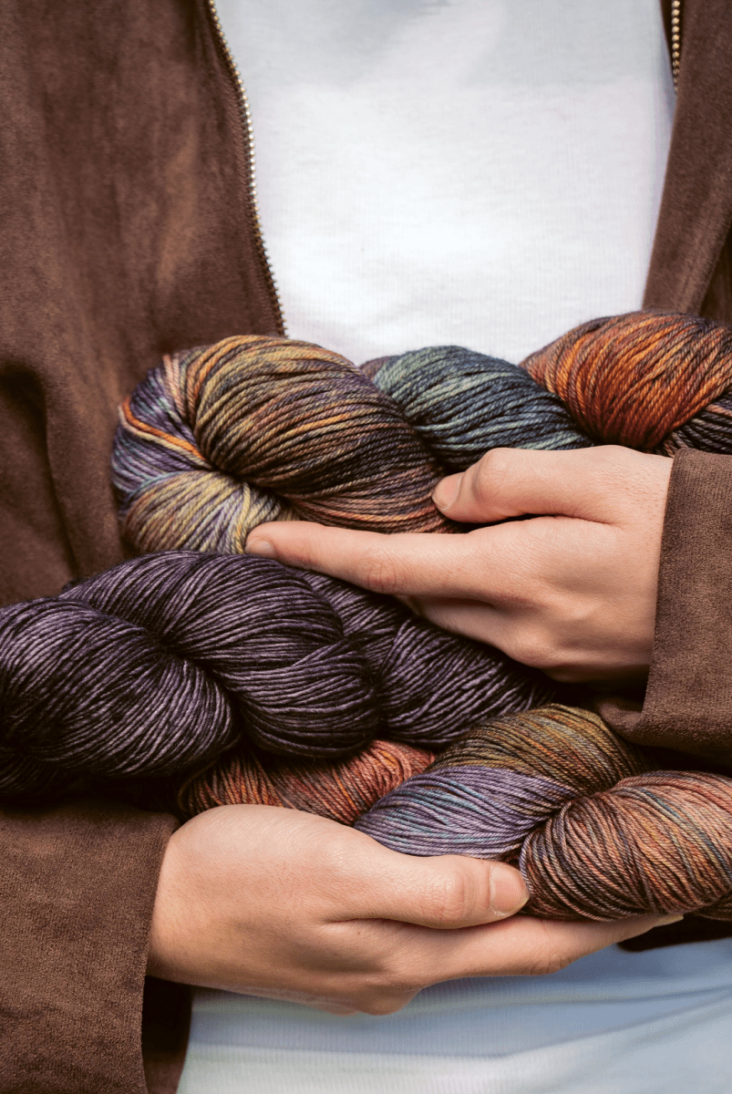 A person wearing a brown jacket and white shirt holds several skeins of Malabrigo Yarn’s Timeless Knits, ideal hand-dyed yarn for crafting unique knitting projects.